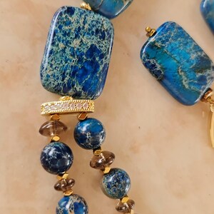 Puede incluir: Un collar con piedras veteadas rectangulares y esf&eacute;ricas en tonos azul y cerceta, intercaladas con cuentas marrones transl&uacute;cidas m&aacute;s peque&ntilde;as y espaciadores dorados. Una pieza conectora dorada rectangular con peque&ntilde;as piedras claras une los segmentos.