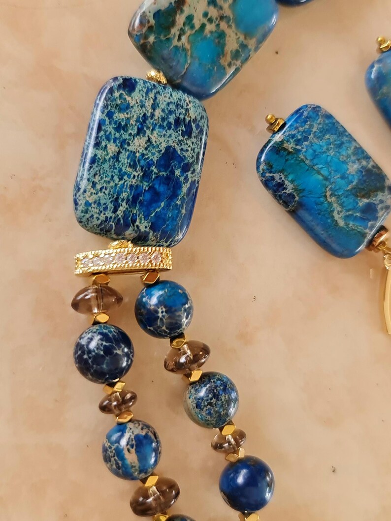Puede incluir: Un collar con piedras veteadas rectangulares y esf&eacute;ricas en tonos azul y cerceta, intercaladas con cuentas marrones transl&uacute;cidas m&aacute;s peque&ntilde;as y espaciadores dorados. Una pieza conectora dorada rectangular con peque&ntilde;as piedras claras une los segmentos.