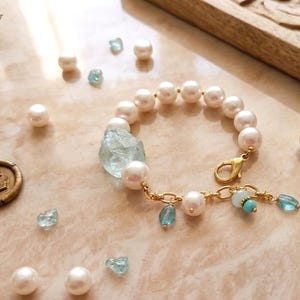 May include: A gold-toned bracelet featuring large, white pearls and a light blue gemstone. The bracelet has a gold clasp and is adorned with smaller blue and turquoise beads. Scattered pearls and gemstones are also visible.