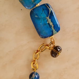 Puede incluir: Un collar con cuentas rectangulares de piedra veteada en azul y turquesa con alambre dorado. Una extensi&oacute;n de cadena dorada cuelga de una cuenta, decorada con una peque&ntilde;a cuenta transl&uacute;cida marr&oacute;n oscuro y una cuenta redonda de piedra azul.