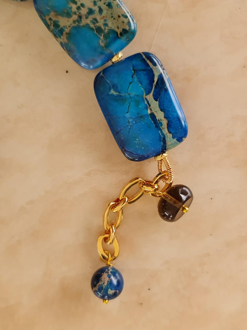 Puede incluir: Un collar con cuentas rectangulares de piedra veteada en azul y turquesa con alambre dorado. Una extensi&oacute;n de cadena dorada cuelga de una cuenta, decorada con una peque&ntilde;a cuenta transl&uacute;cida marr&oacute;n oscuro y una cuenta redonda de piedra azul.
