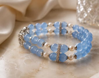 Puede incluir: Una pulsera de doble hilo con cuentas azul claro translúcidas y pequeñas perlas blancas. El diseño incluye espaciadores de metal plateado y dos barras verticales incrustadas con cristales transparentes, unidas por un cierre decorativo.