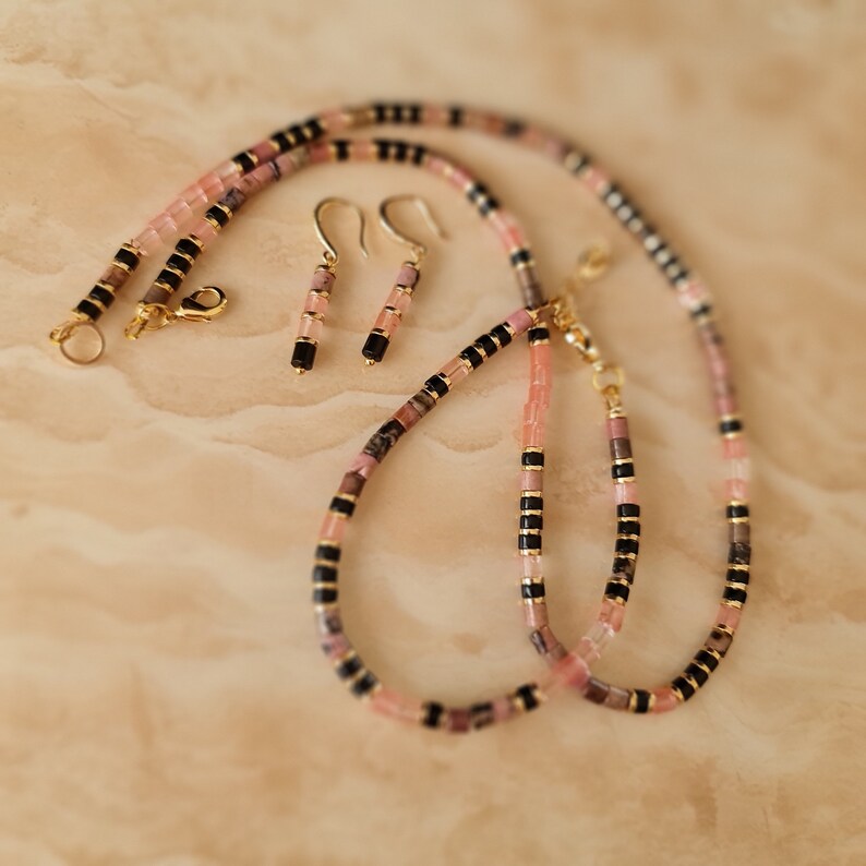 May include: A pink and black beaded necklace and matching earrings. The necklace is made of small, rectangular beads in shades of pink and black. The earrings are simple and dangle.