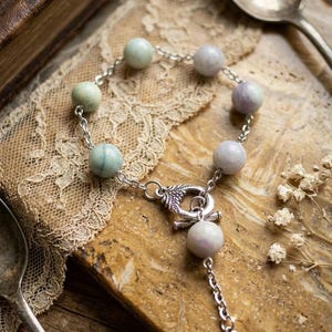 May include: A beaded bracelet featuring round, pastel-colored gemstones strung on a silver chain. The bracelet has a decorative clasp and a dangling charm with a small heart. The background includes a vintage book, lace, and antique silverware.