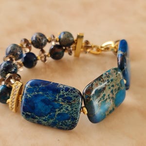 Puede incluir: Una pulsera con grandes cuentas rectangulares de piedra moteada azul y verde, cuentas de piedra azul redondas m&aacute;s peque&ntilde;as y cuentas marrones transl&uacute;cidas. El dise&ntilde;o incluye espaciadores de metal dorado, un cierre de barra dorado y una peque&ntilde;a pieza de acento dorada.