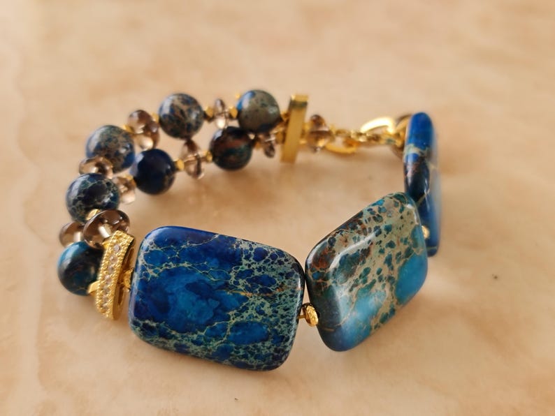 Puede incluir: Una pulsera con grandes cuentas rectangulares de piedra moteada azul y verde, cuentas de piedra azul redondas m&aacute;s peque&ntilde;as y cuentas marrones transl&uacute;cidas. El dise&ntilde;o incluye espaciadores de metal dorado, un cierre de barra dorado y una peque&ntilde;a pieza de acento dorada.