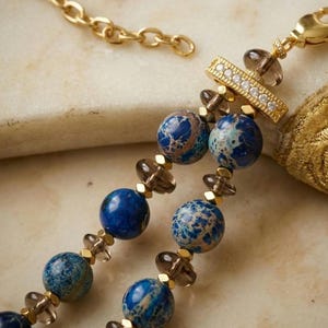 Puede incluir: Un collar de doble hebra con cuentas redondas jaspeadas en azul y marr&oacute;n, cuentas transl&uacute;cidas de color gris humo y peque&ntilde;os espaciadores dorados. Una barra rectangular dorada con una fila de piedras claras une las hebras a una cadena.