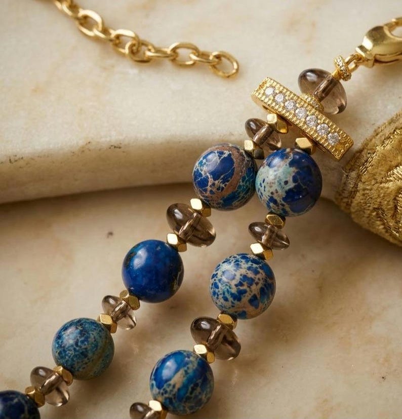 Puede incluir: Un collar de doble hebra con cuentas redondas jaspeadas en azul y marr&oacute;n, cuentas transl&uacute;cidas de color gris humo y peque&ntilde;os espaciadores dorados. Una barra rectangular dorada con una fila de piedras claras une las hebras a una cadena.