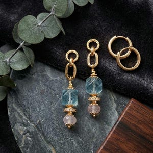 May include: Gold-toned earrings with blue and pink gemstones. The earrings feature a chain design with a large, light blue gemstone at the top and a smaller, pink gemstone at the bottom. A pair of gold hoop earrings are also present.