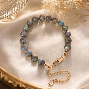 May include: A beaded bracelet featuring faceted, round, grey-toned labradorite stones with subtle iridescent flashes. The bracelet includes a gold-toned metal lobster claw clasp and an adjustable chain extension finished with a small heart-shaped charm.