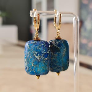 Puede incluir: Un par de pendientes colgantes con cuentas rectangulares de piedra veteada en azul y turquesa, suspendidas de cierres de palanca de metal dorado. Cada piedra muestra un patr&oacute;n &uacute;nico de vetas azul oscuro y turquesa con detalles met&aacute;licos.