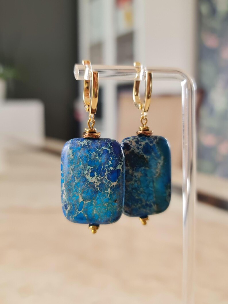 Puede incluir: Un par de pendientes colgantes con cuentas rectangulares de piedra veteada en azul y turquesa, suspendidas de cierres de palanca de metal dorado. Cada piedra muestra un patr&oacute;n &uacute;nico de vetas azul oscuro y turquesa con detalles met&aacute;licos.