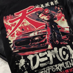 Könnte beinhalten: Schwarzes T-Shirt mit einem rot-weißen Grafikdesign, das ein Auto und eine Figur zeigt. Das Design enthält japanischen Text und das Wort "DEMON" in großen weißen Buchstaben. Der Stil ist von Anime und Street Racing inspiriert.