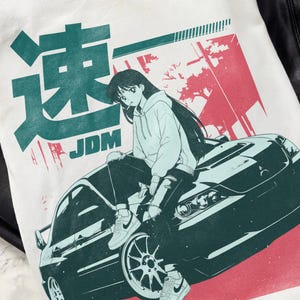 May include: White t-shirt featuring a teal and red graphic. The graphic includes Japanese characters, the word "JDM", and an illustration of a person sitting on a car. The person is wearing a hoodie and sneakers.