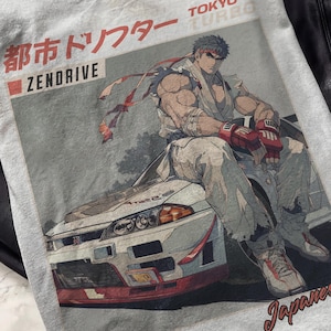 May include: A light gray t-shirt featuring a graphic of a muscular anime character sitting on a white and red sports car. The shirt has Japanese text and the words "Zendrive" and "Tokyo Turbo". The design is in shades of gray, red, and white.