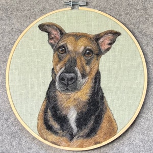 May include: A needle felted portrait of a brown and black dog with a white patch on its chest. The dog is looking directly at the viewer with a serious expression. The portrait is framed in a wooden hoop.