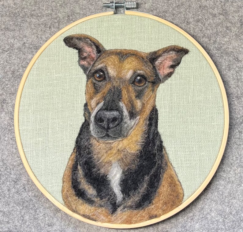 May include: A needle felted portrait of a brown and black dog with a white patch on its chest. The dog is looking directly at the viewer with a serious expression. The portrait is framed in a wooden hoop.