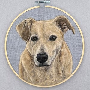May include: A needle felted portrait of a dog in a wooden embroidery hoop. The dog is light brown with a black nose and dark eyes. The background is a light gray fabric.