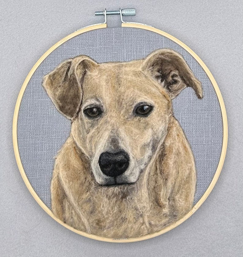 May include: A needle felted portrait of a dog in a wooden embroidery hoop. The dog is light brown with a black nose and dark eyes. The background is a light gray fabric.