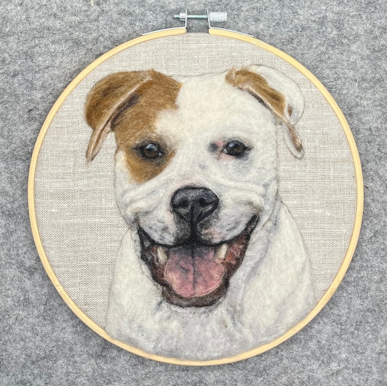 May include: A needle felted portrait of a white and brown dog with a happy expression. The dog is smiling with its tongue sticking out. The portrait is framed in a wooden hoop.