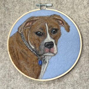 May include: A brown and white dog with a blue collar is depicted in needle felting on a light blue background. The dog is framed in a wooden hoop.