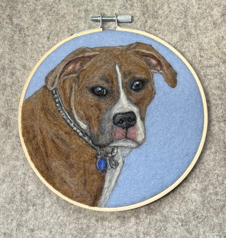 May include: A brown and white dog with a blue collar is depicted in needle felting on a light blue background. The dog is framed in a wooden hoop.