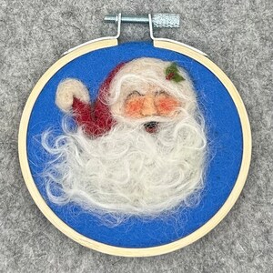 May include: A needle felted Santa Claus head with a white beard and red hat, framed in a wooden hoop with a blue background.
