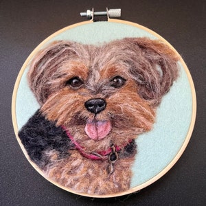 May include: A needle felted Yorkshire Terrier dog portrait in a wooden embroidery hoop. The dog is brown and black with a pink tongue sticking out. The background is light blue.