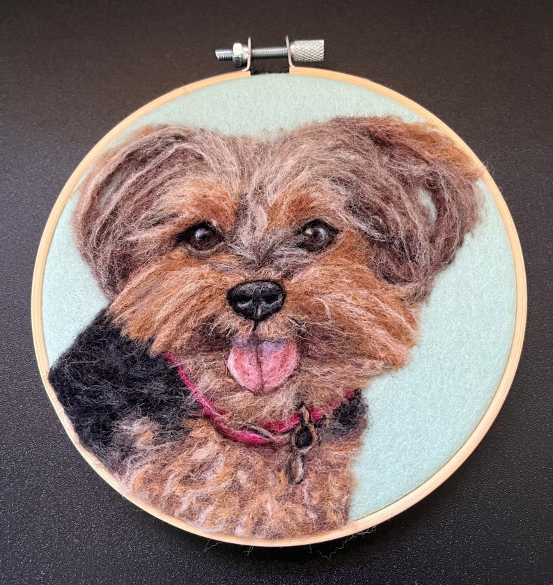May include: A needle felted Yorkshire Terrier dog portrait in a wooden embroidery hoop. The dog is brown and black with a pink tongue sticking out. The background is light blue.