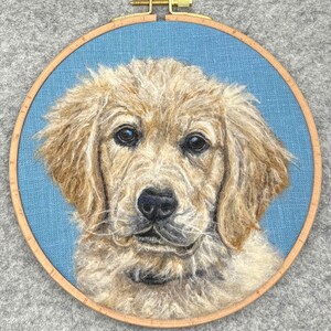 May include: A needle felted portrait of a golden retriever puppy in a wooden embroidery hoop. The puppy has a light brown coat and a sweet expression.