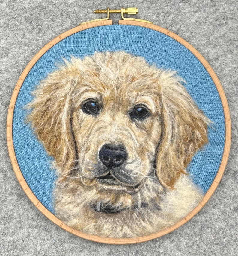May include: A needle felted portrait of a golden retriever puppy in a wooden embroidery hoop. The puppy has a light brown coat and a sweet expression.