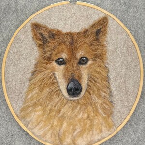 May include: A close-up of a needle felted portrait of a dog. The dog has brown fur and a black nose. The portrait is framed in a wooden hoop.