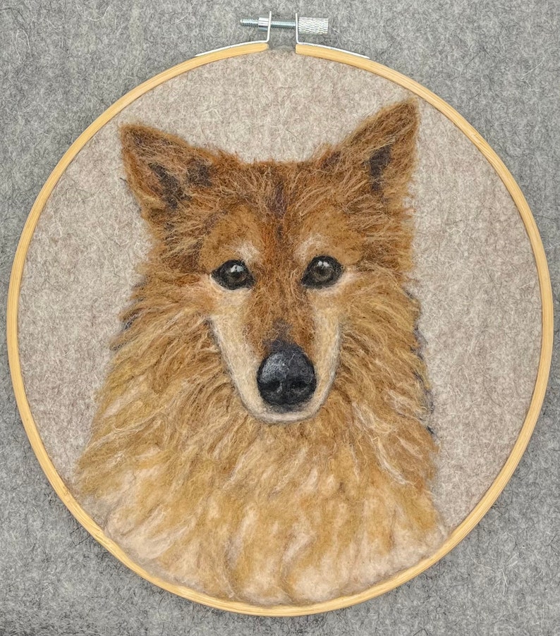 May include: A close-up of a needle felted portrait of a dog. The dog has brown fur and a black nose. The portrait is framed in a wooden hoop.