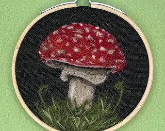 May include: A needle felted mushroom with a red cap with white spots and a white stem. The mushroom is surrounded by green felt grass and is framed in a wooden hoop.