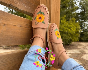 Könnte beinhalten: Ein Paar braune Ledersandalen mit Sonnenblumen-Stickerei. Die Sandalen haben Knöchelriemen und werden an den Füßen einer Person getragen. Die Sandalen liegen auf einem Holzzaun.