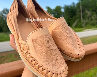 Könnte beinhalten: Ein Paar hellbraune Leder-Huarache-Ballerinas mit floraler Prägung. Die Schuhe haben geflochtene Details um die Zehen und eine flache Sohle. Der Text "Ruby Tradicion En Moda" ist sichtbar.