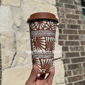 May include: A brown ceramic to-go cup with a white lid and a white floral design. The cup has the text "Ruby Tradicion En Moda" on the side.
