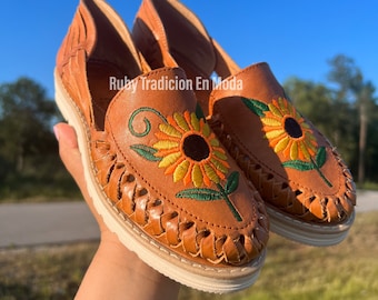 Könnte beinhalten: Ein Paar braune Ledersandalen mit einem gestickten Sonnenblumenmuster. Die Sandalen haben einen geflochtenen Lederriemen und eine weiße Sohle.