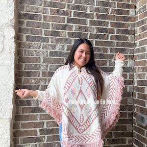 Peut inclure: Un poncho blanc avec des franges roses et un motif géométrique coloré. Le poncho a un col en V et des manches longues. Le texte "Ruby Tradicion En Moda" est visible sur le poncho.
