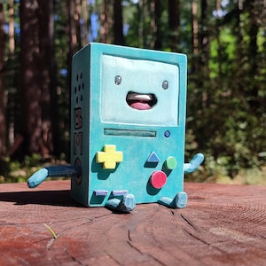 Puede incluir: Un robot de juguete de madera azul verdoso con una cara sonriente y una pantalla que dice "BMO". El robot tiene botones amarillos, verdes y rojos.