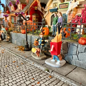 May include: A miniature Halloween village scene with cobblestone streets, featuring decorative houses, pumpkins, and costumed figures. A firefighter figure holds a jack-o'-lantern, and a person in a french fries costume walks down the street. A ghost and the words "Trick or Treat" are visible.