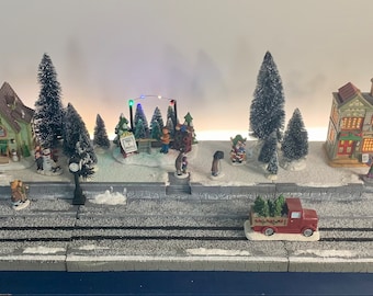 May include: A miniature Christmas village scene with snow-covered houses, trees, and figurines. A red truck carries Christmas trees. The scene includes a street, sidewalks, and a snowy landscape, creating a festive holiday display.