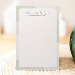 Könnte beinhalten: Rechteckiger Notizblock mit weißem Innenraum und hellgrünem Rand. Der Text "Hannah's Notizen" ist oben in eleganter Schreibschrift geschrieben. Der Notizblock hat ein minimalistisches Design, geeignet für Notizen.