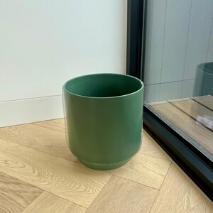 May include: A green ceramic planter with a smooth, matte finish. The planter is sitting on a light wood floor with a herringbone pattern.