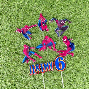 May include: A set of 7 cake toppers featuring Spiderman in various poses, a blue number 6, and the name "JEREMY" in red and yellow letters. The toppers are on sticks and are set against a green grass background.