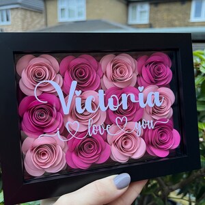 May include: A black framed shadow box with a white background and pink paper flowers. The text "Victoria I love you" is written in white on the background.