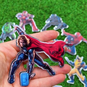 Puede incluir: Una colecci&oacute;n de pegatinas de personajes de superh&eacute;roes, incluyendo a Thor con su martillo y capa roja, y otros personajes de Marvel. Las pegatinas est&aacute;n sobre un fondo de hierba verde.