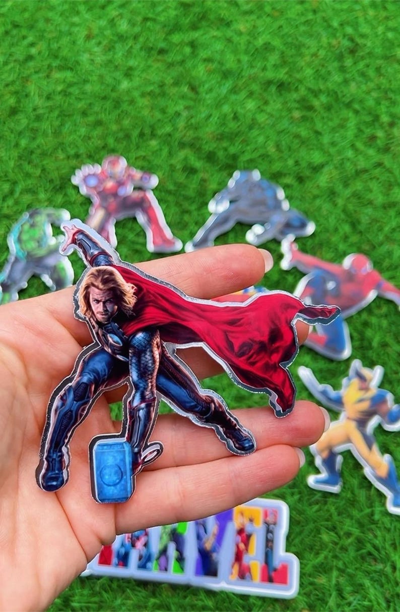 Puede incluir: Una colecci&oacute;n de pegatinas de personajes de superh&eacute;roes, incluyendo a Thor con su martillo y capa roja, y otros personajes de Marvel. Las pegatinas est&aacute;n sobre un fondo de hierba verde.