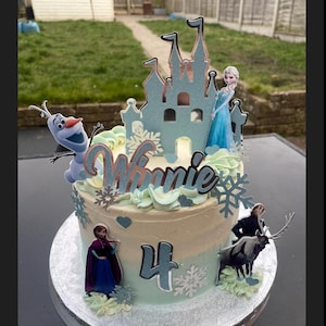 Op de afbeelding: Een lichtblauwe en witte geglazuurde taart versierd met een Frozen-thema. De taart heeft een kasteel, Olaf, Elsa, Anna en Sven-figuurtjes. De taart is versierd met witte sneeuwvlokken en het nummer 4. De taart zegt "Winnie" op de bovenkant.