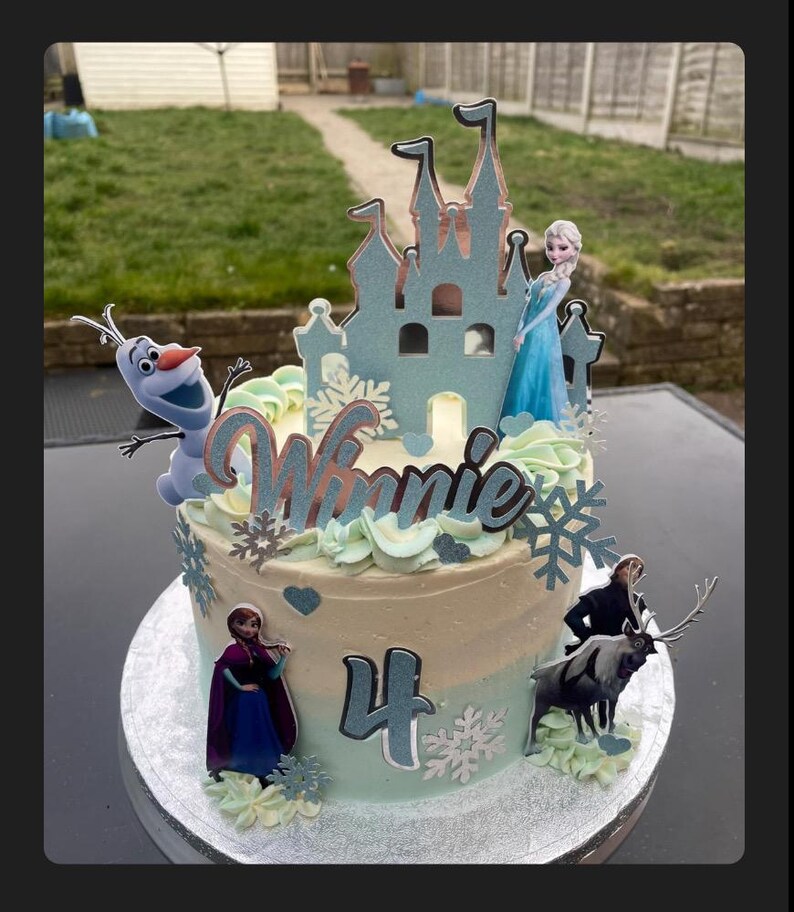 Op de afbeelding: Een lichtblauwe en witte geglazuurde taart versierd met een Frozen-thema. De taart heeft een kasteel, Olaf, Elsa, Anna en Sven-figuurtjes. De taart is versierd met witte sneeuwvlokken en het nummer 4. De taart zegt "Winnie" op de bovenkant.